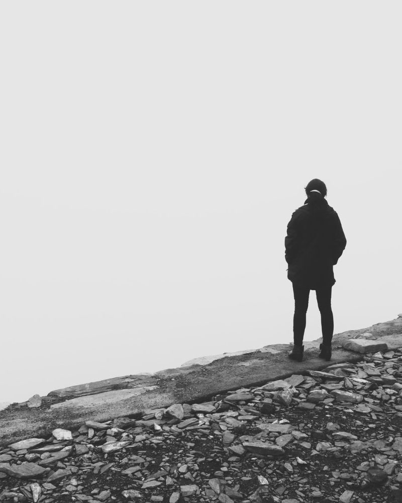 grey-scale-shot-person-standing-edge-cliff-looking-white-void-min